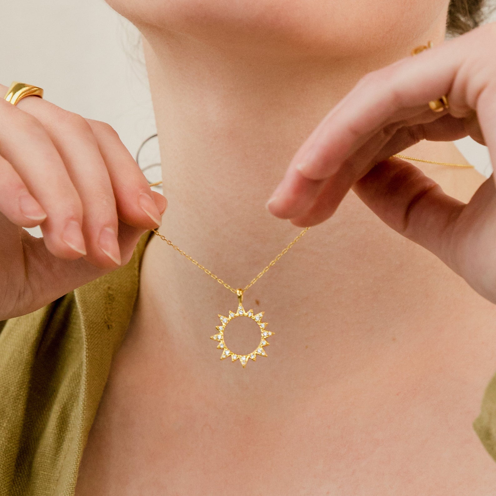 A person holds the Pave Sun Necklace, featuring a gold sun-shaped pendant, near their neck while wearing gold rings and a green top.