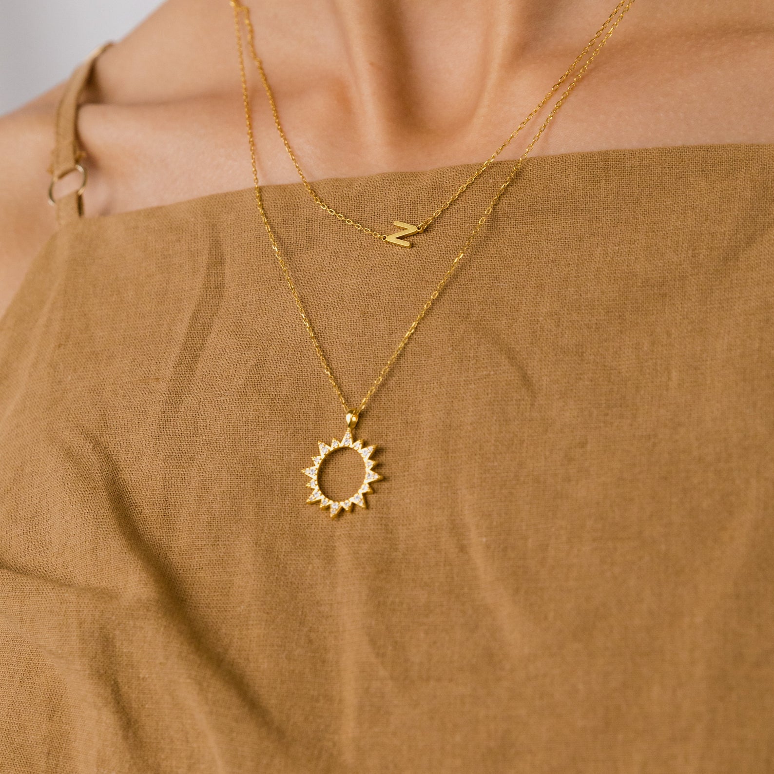 Woman in a brown dress wears two gold necklaces: the Pave Sun Necklace, featuring a sun pendant with pave accents, and another adorned with the letter N.