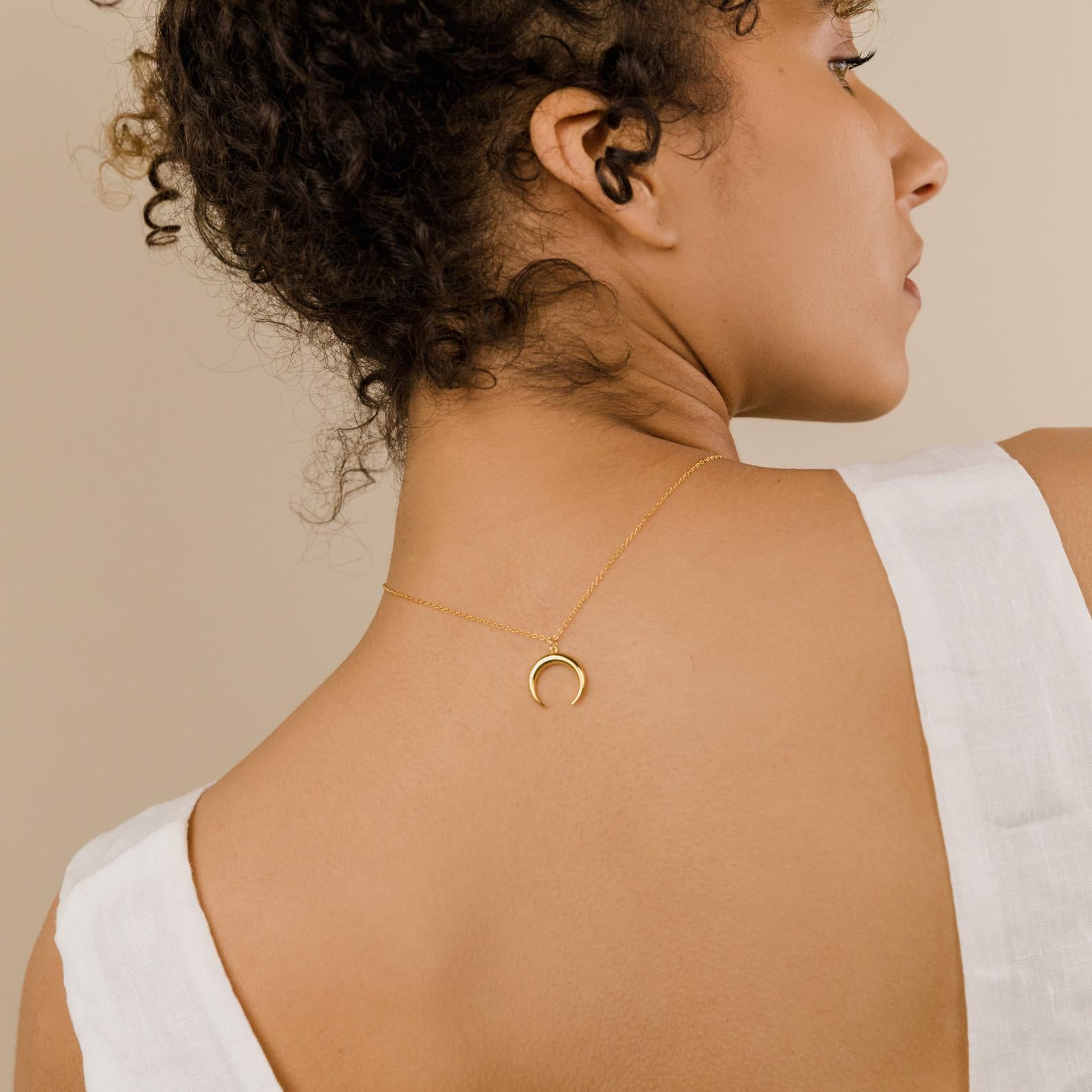 A woman with curly hair wears the Crescent Moon Necklace backwards, showcasing its elegant amulet on her bare back while dressed in white.
