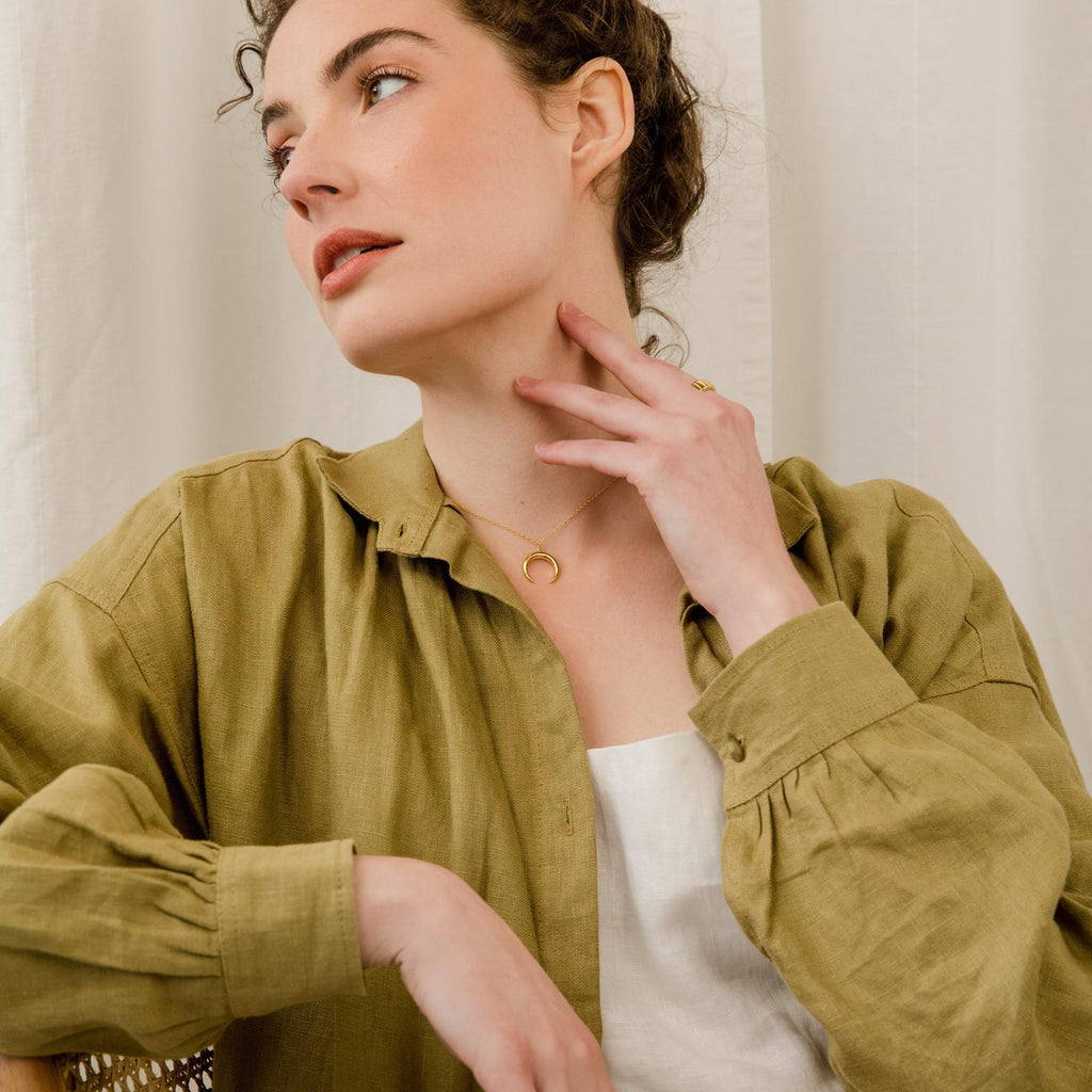 A woman in an olive green shirt touches her neck and looks to the side, showcasing the Crescent Moon Necklace.