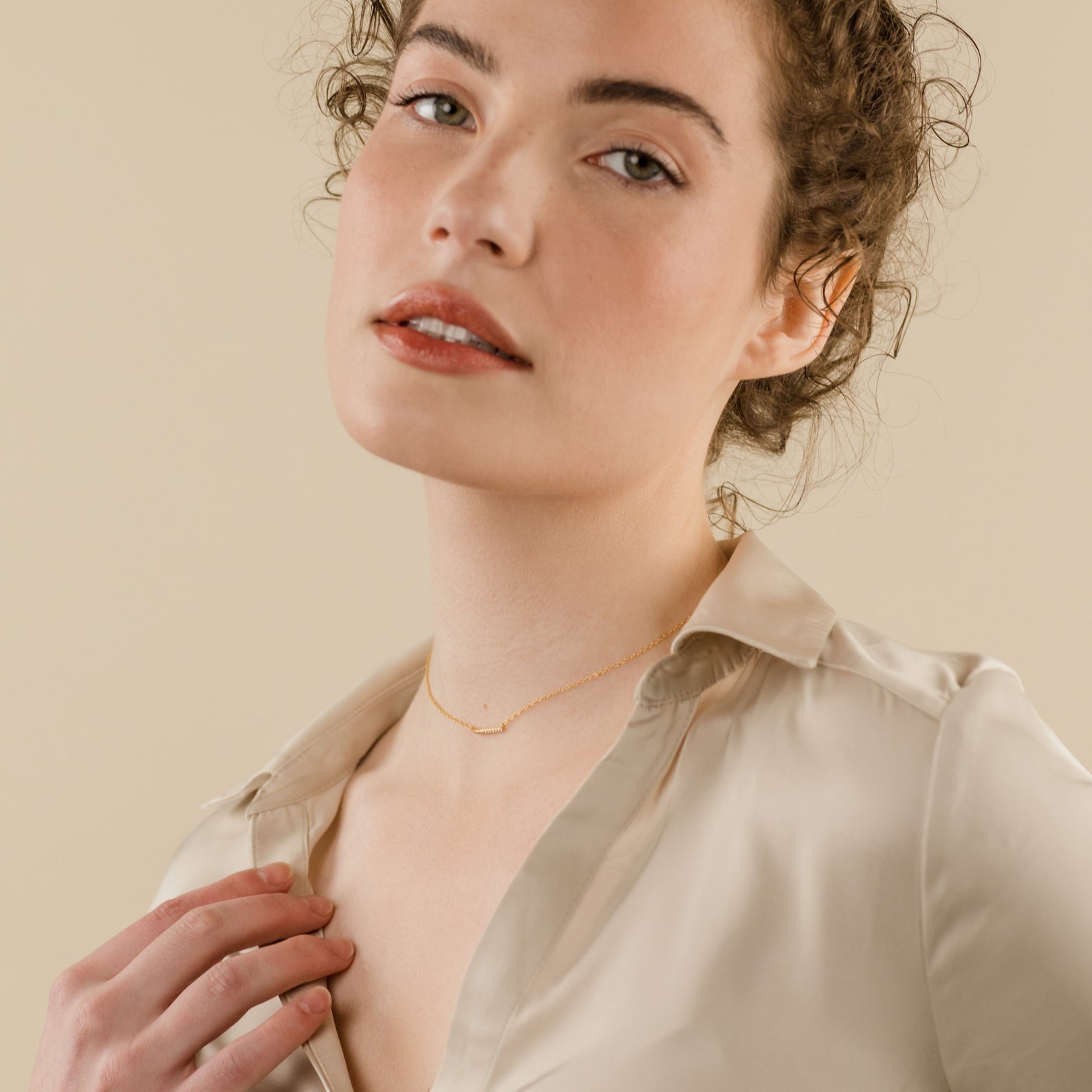 A woman with curly hair in a beige blouse poses against a neutral background, highlighting the Pave Bar Necklace in a delicate gold stack.