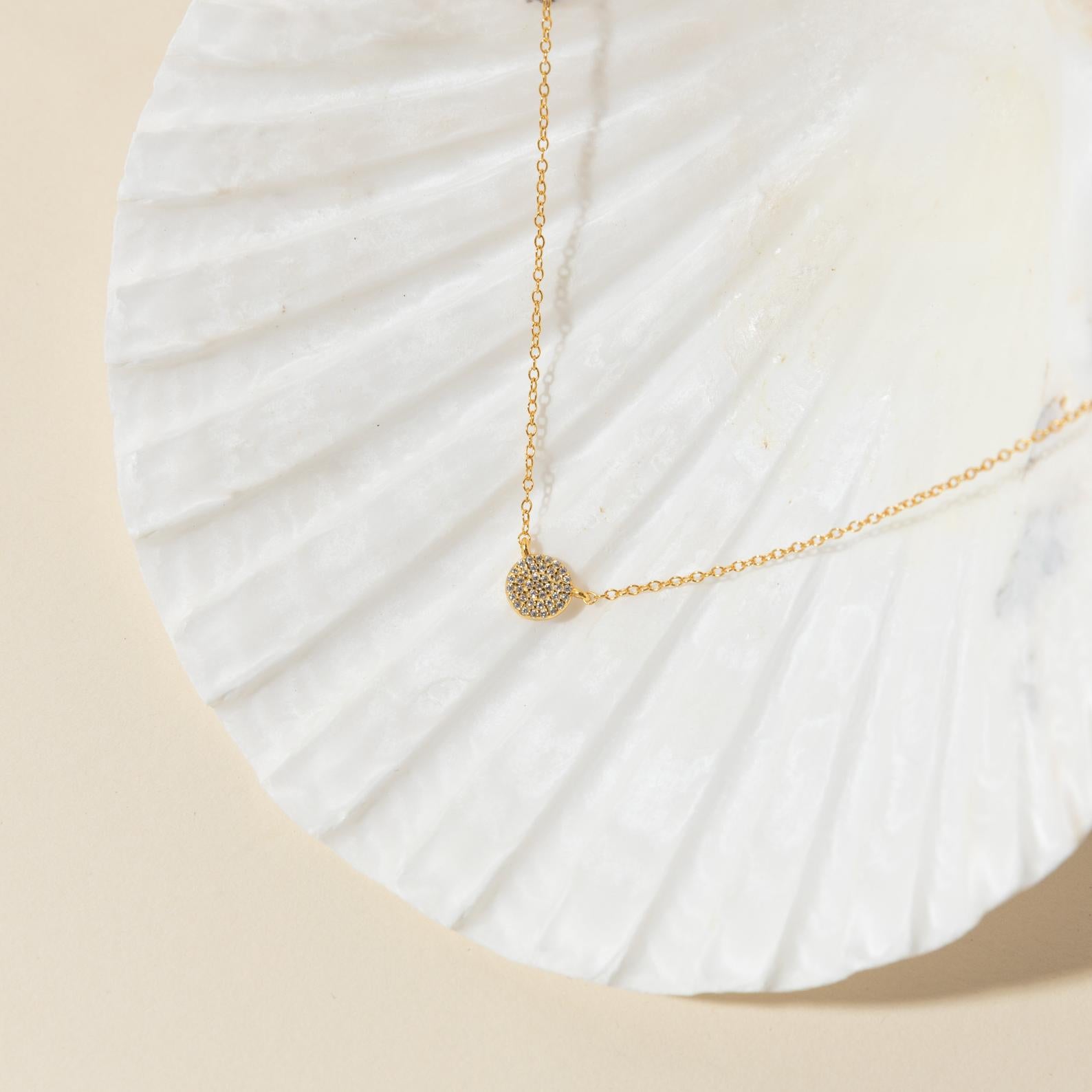 The Pave Round Necklace with a silver chain is displayed on a white seashell against a beige background.