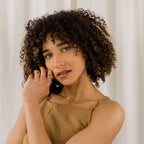 A woman with curly hair, gold jewelry, and a tan dress poses in front of a white curtain, showcasing the Pave Round Necklace.