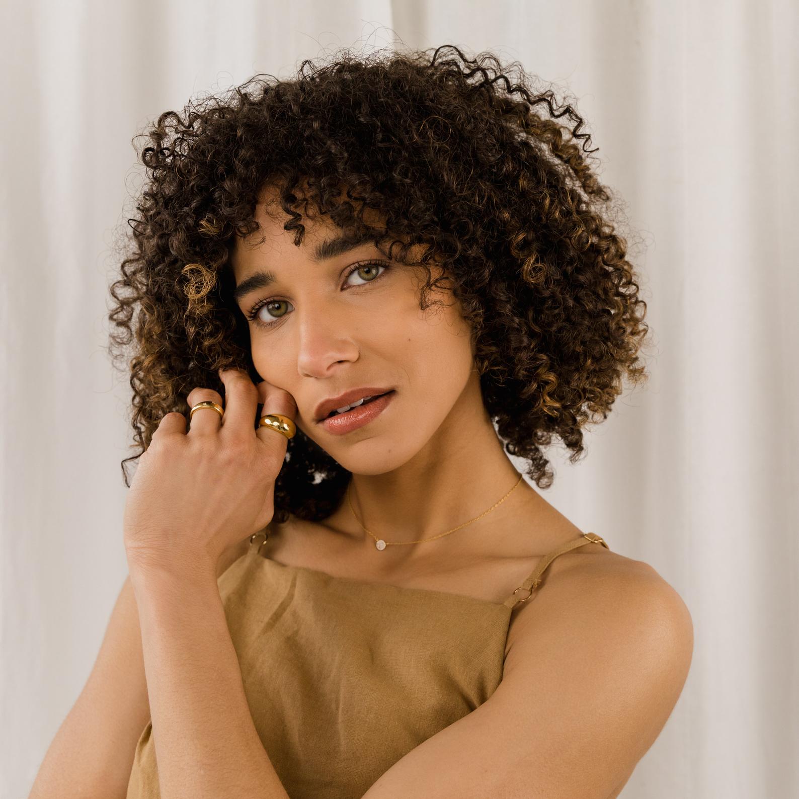 A woman with curly hair, gold jewelry, and a tan dress poses in front of a white curtain, showcasing the Pave Round Necklace.