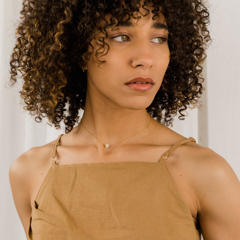 A woman with curly hair in a tan sleeveless top looks to the side, wearing the Pave Round Necklace—a delicate round pendant on a silver chain.