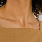 Gold necklace with a delicate round pendant encrusted in tiny pave set stones, worn just below the collarbone with a square neckline in a neutral linen fabric.