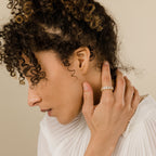 A woman with curly hair gently touches her neck, showcasing the Alannah Ring—an oval diamond accent—paired with a white pleated top for an effortlessly elegant look.
