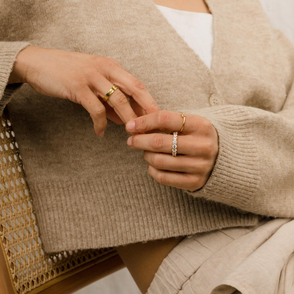 Close-up of a person wearing the Christy Ring along with other feminine jewelry and a beige sweater, sitting on a woven chair.