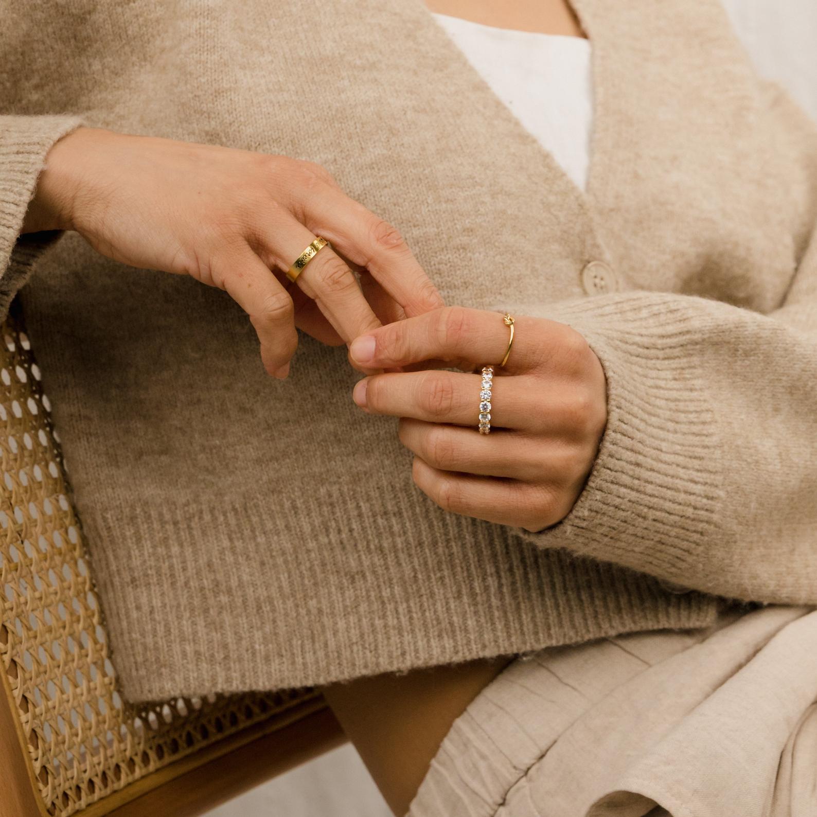 Close-up of a person wearing the Christy Ring along with other feminine jewelry and a beige sweater, sitting on a woven chair.