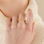A hand wearing the Christy Ring—a gold stacking ring with clear gemstones—rests on a cream knit turtleneck sweater.