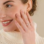 A smiling woman touches her face, wearing a cream turtleneck and showcasing the elegant Christy Ring on her finger.