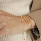 A wrist styled with two delicate gold bracelets, including the Constellation Bracelet, paired with a beige ribbed top and tan blazer sleeve.