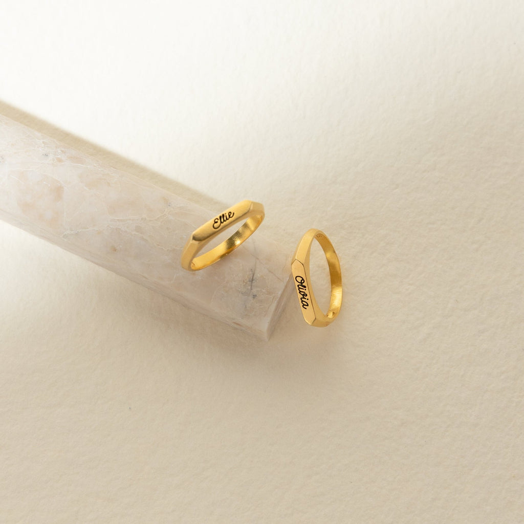 Two Paris Thin Name Signet Rings engraved with “Elle” rest on a light stone and cream backdrop—ideal for stacking or as a timeless statement piece.