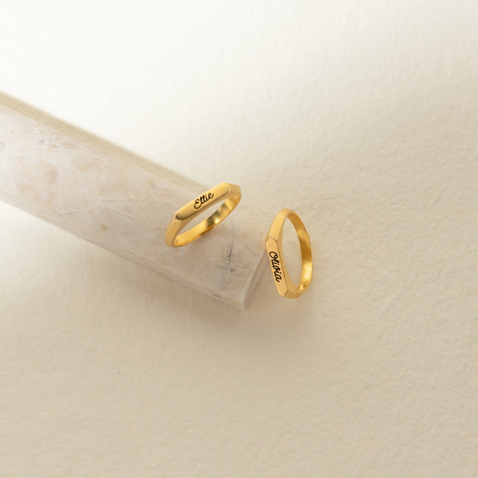 Two Paris Thin Name Signet Rings engraved with “Elle” rest on a light stone and cream backdrop—ideal for stacking or as a timeless statement piece.