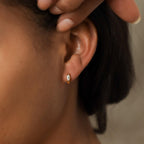 A close-up of a person’s ear wearing Aurora Huggies—small gold hoops featuring a marquise-cut gem and delicate diamond accents.