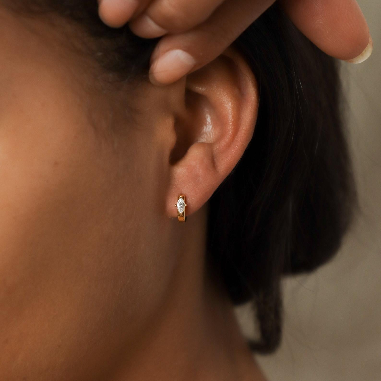 A close-up of a person’s ear wearing Aurora Huggies—small gold hoops featuring a marquise-cut gem and delicate diamond accents.
