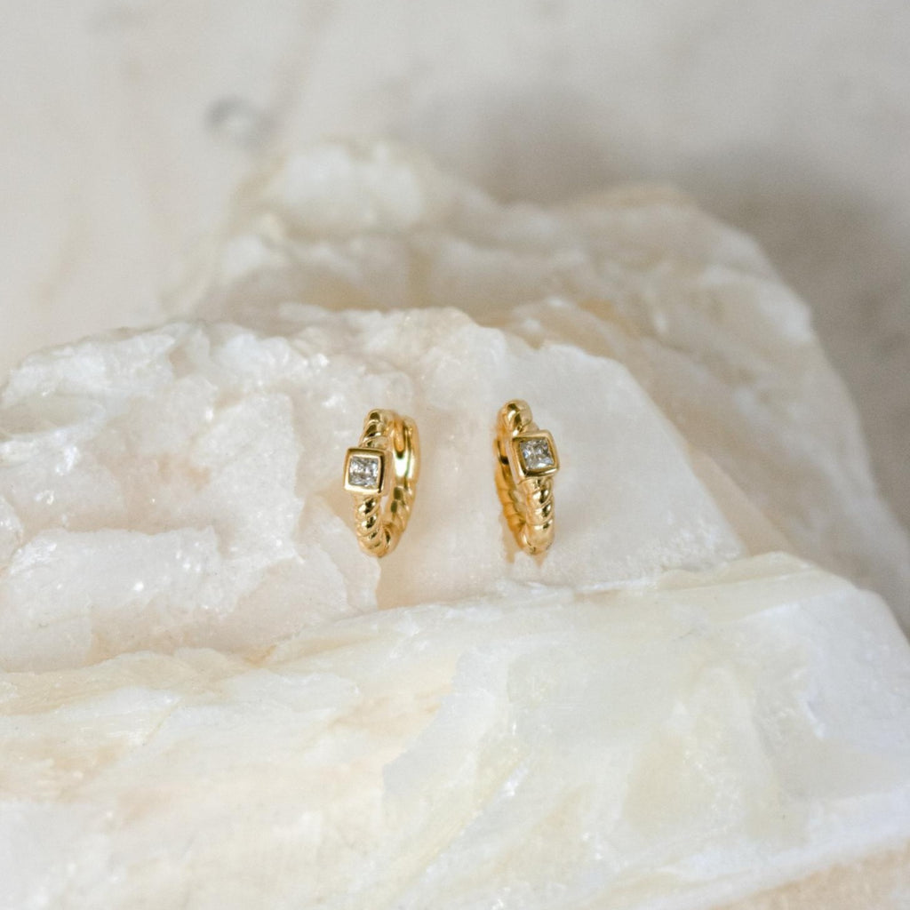 Willow Huggies: Gold hoop earrings with square, clear stones set in 925 sterling silver, shown on a textured white stone surface.