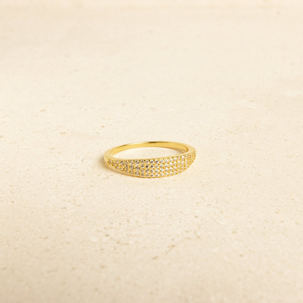 The Gina Pave Signet Ring, a gold statement piece adorned with small sparkling stones, is displayed on a light beige surface.