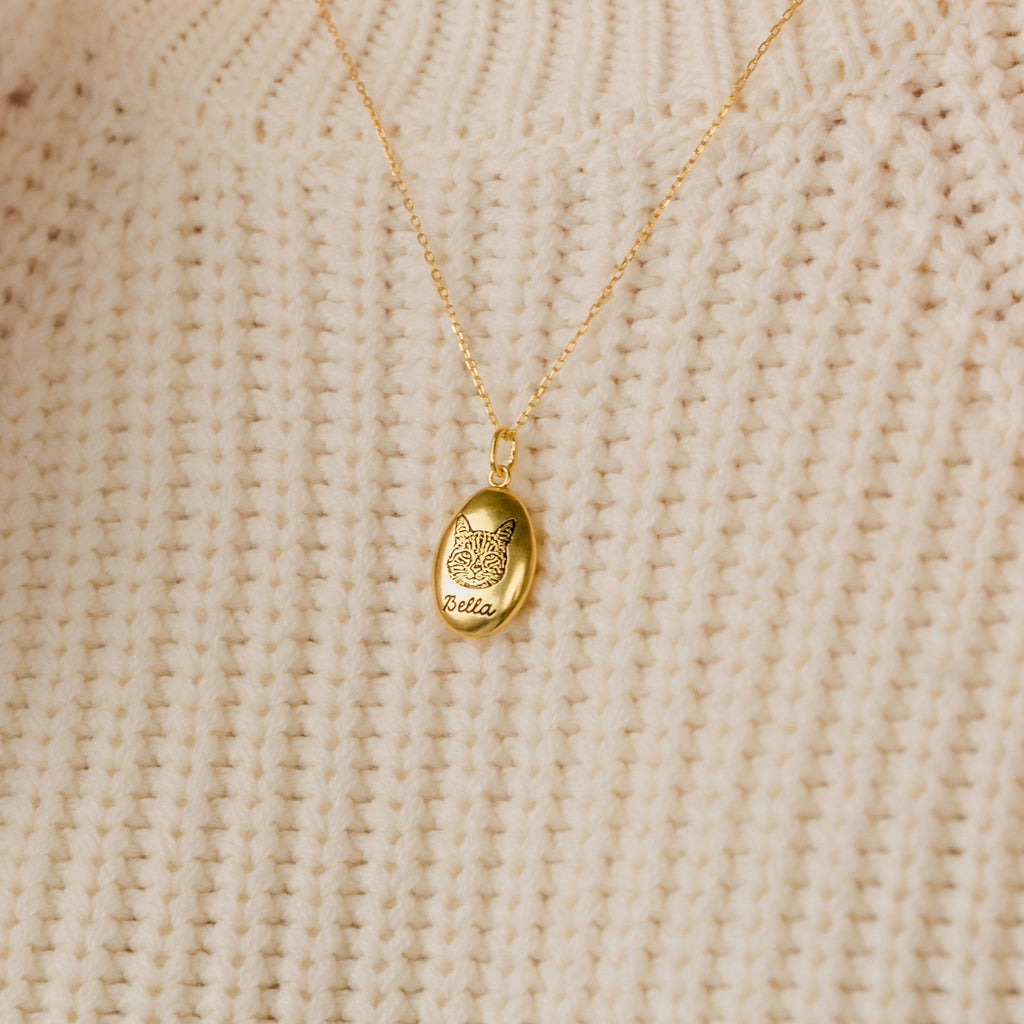 The Oval Pet Necklace, engraved with a butterfly and the name Bella, rests elegantly on a cream knit sweater—ideal for anyone who loves personalized pet jewelry.