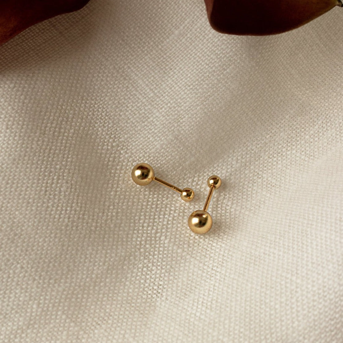 The Hailey Ball Studs, featuring a gold finish and secure screw-on backing, are displayed on a textured cream fabric background.