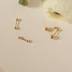 Two pairs of Hailey Ball Studs in gold (3mm and 5mm) with secure screw-on backs rest on a cream surface beside white flowers—ideal as stylish starter earrings.