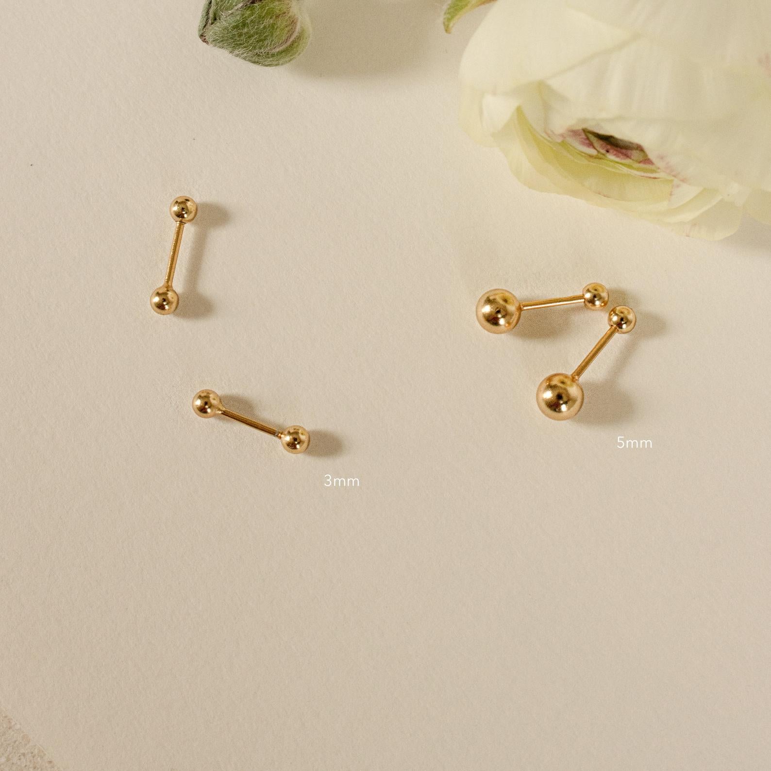 Two pairs of Hailey Ball Studs in gold (3mm and 5mm) with secure screw-on backs rest on a cream surface beside white flowers—ideal as stylish starter earrings.