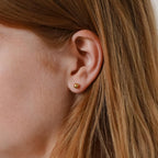 A close-up of a woman's ear wearing Hailey Ball Studs—small gold ball earrings with secure screw-on backs. Light brown hair is partially visible, making these studs an ideal choice for comfortable starter earrings.
