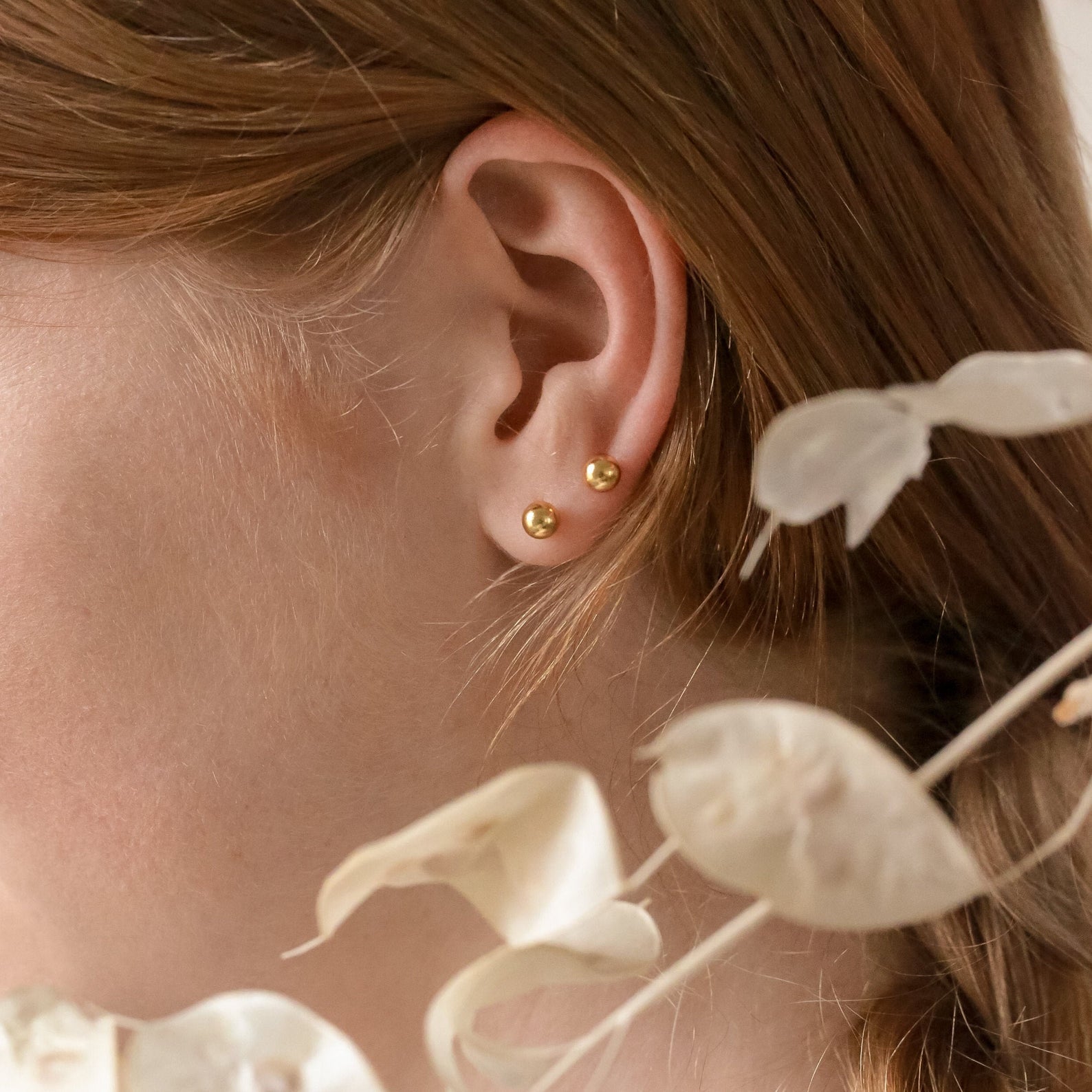 Close-up of a woman’s ear wearing Hailey Ball Studs—gold starter earrings with screw-on backs—accented by dried white flowers in the foreground.