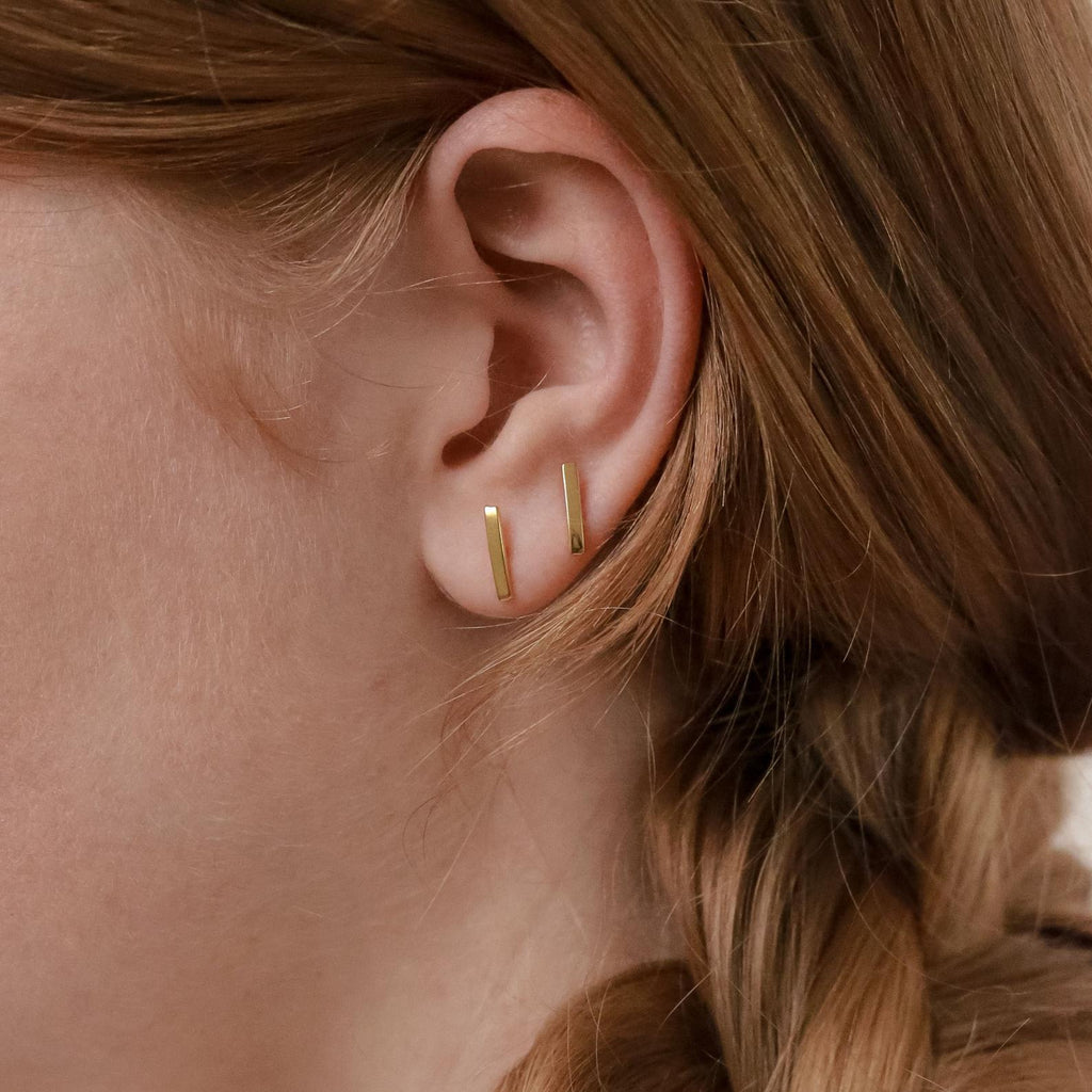 Close-up of a person’s ear wearing the Dainty Bar Studs, shown as two small vertical gold earrings, with auburn braided hair.