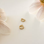 The Esther Duo Pave Huggies, a pair of gold double hoop earrings adorned with small crystals, are displayed on a white surface near light pink flowers.