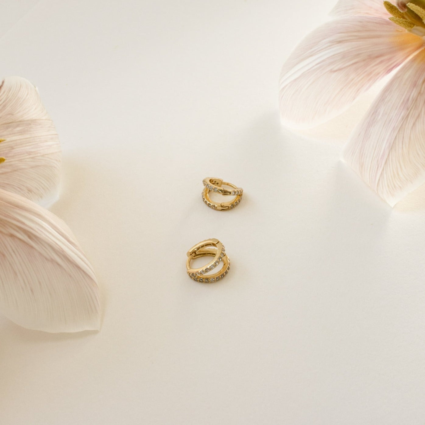 The Esther Duo Pave Huggies, a pair of gold double hoop earrings adorned with small crystals, are displayed on a white surface near light pink flowers.
