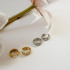 Morgan Croissant Hoops in gold and silver, inspired by the French girl aesthetic, are displayed on a white surface beside pale flowers.