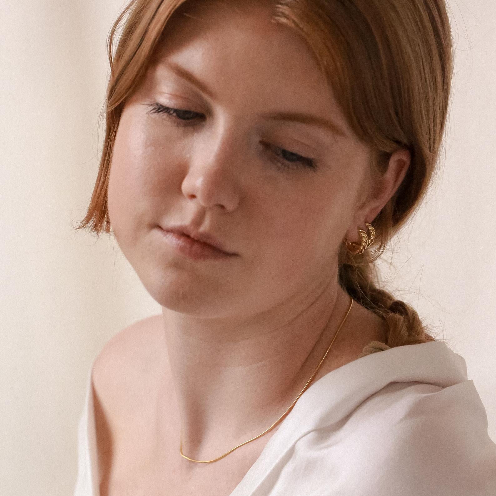 A red-haired woman looks down, wearing the Morgan Croissant Hoops and a gold necklace with a white top, creating an effortless French girl aesthetic.
