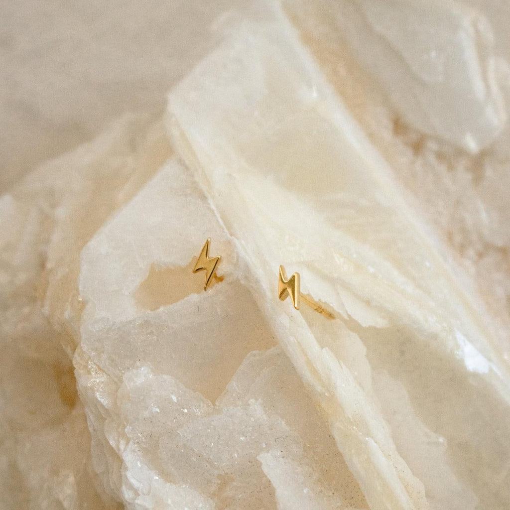 Lightning Bolt Studs in gold are displayed on a textured pale stone surface, making them a striking, edgy addition to your jewelry collection.