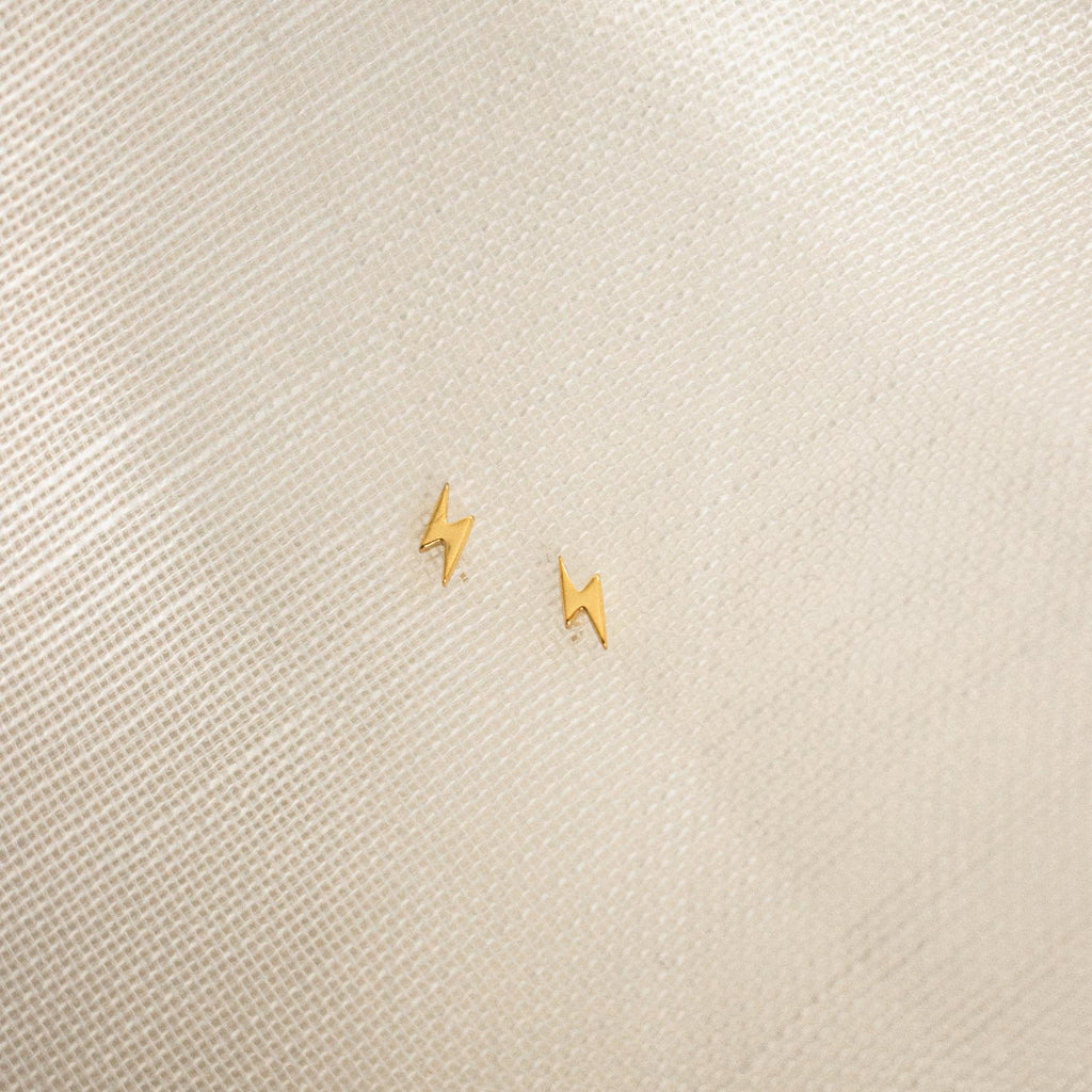 Lightning Bolt Studs in small gold rest on a textured light beige fabric background, adding a subtle yet edgy touch to any outfit.