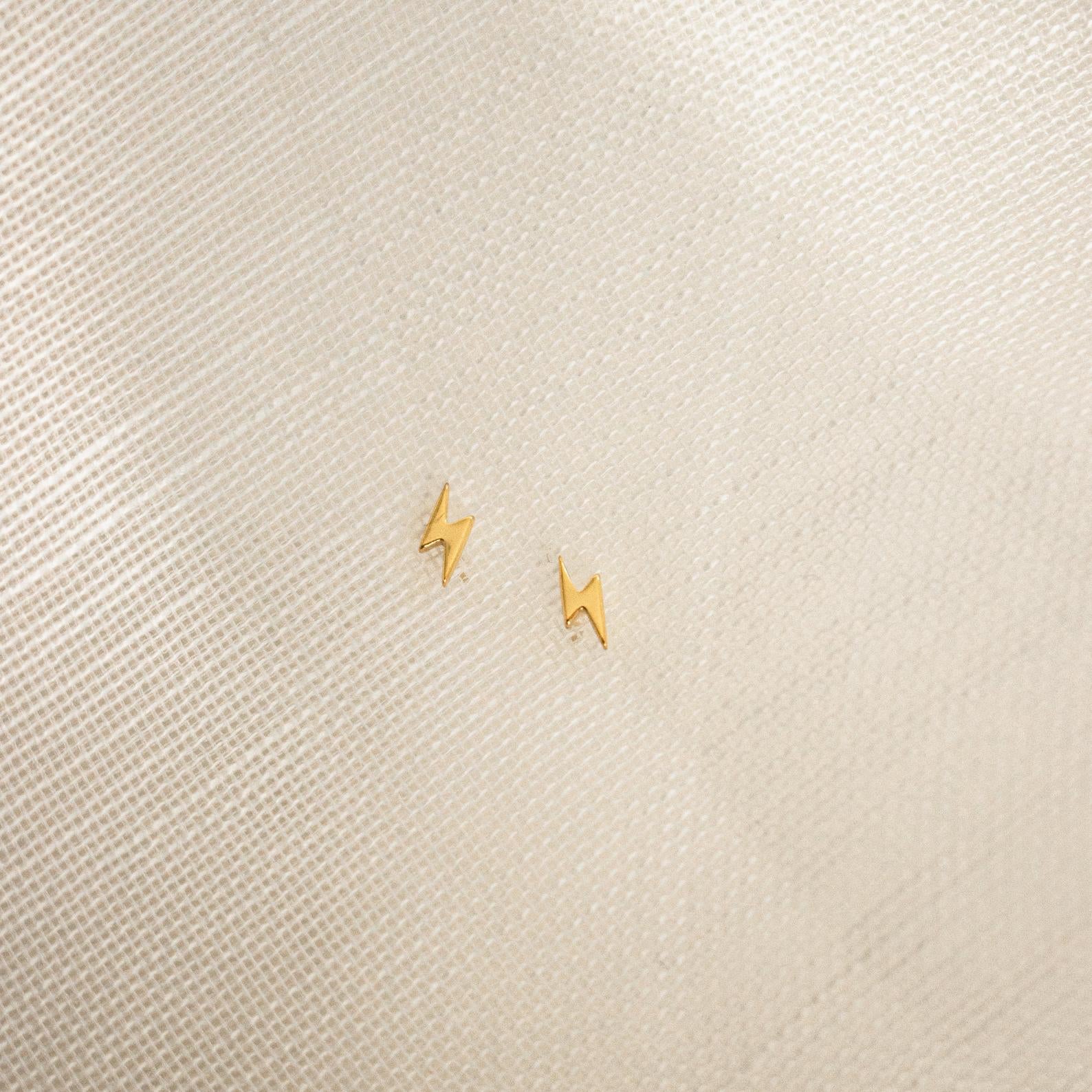 Lightning Bolt Studs in small gold rest on a textured light beige fabric background, adding a subtle yet edgy touch to any outfit.