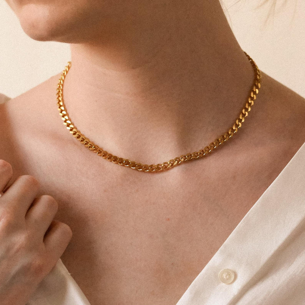 A gold curb chain necklace worn around a woman’s neck with an off-shoulder cream top.
