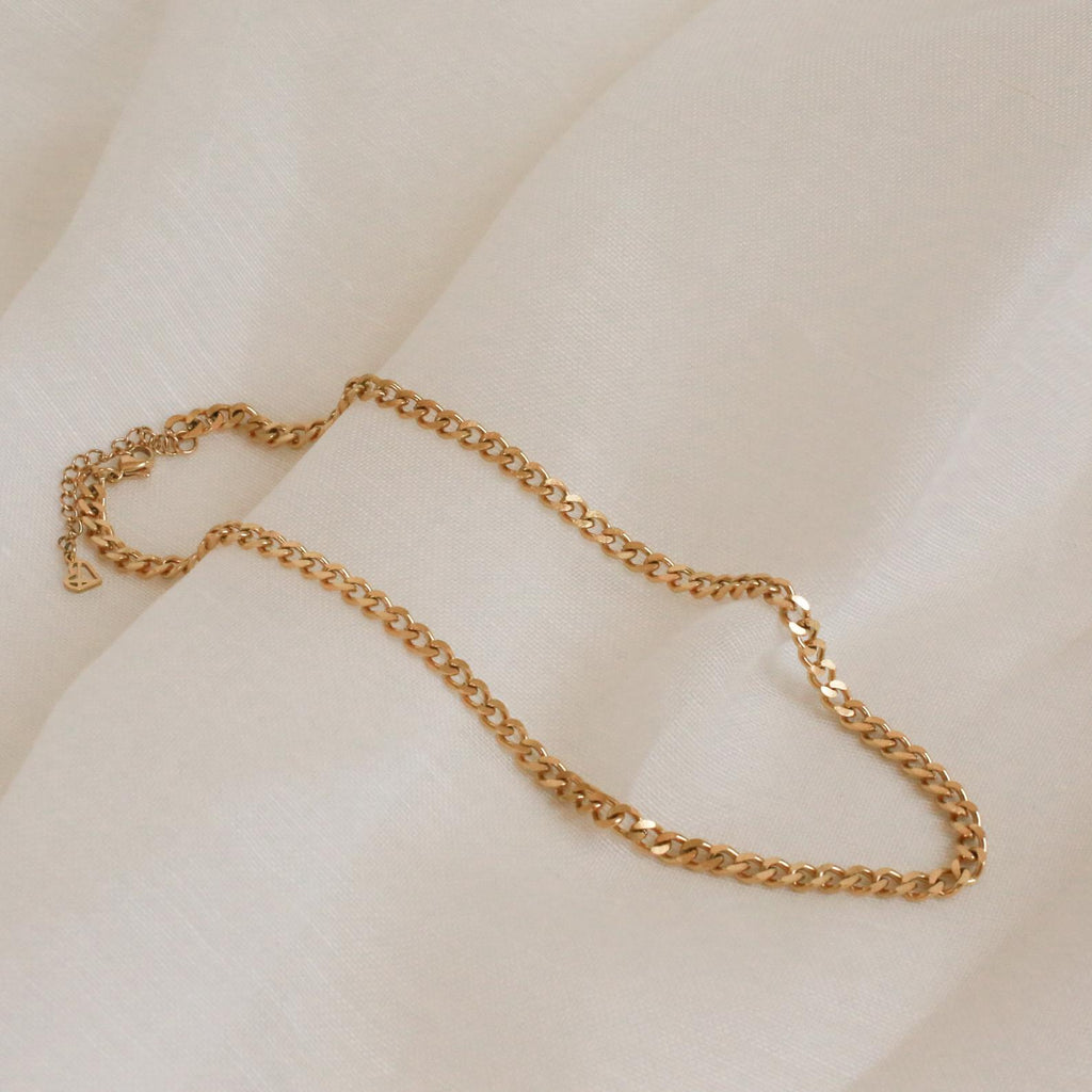 A close-up shot of a chunky gold curb chain necklace with flat, interlocking links, draped gently across a soft ivory-colored fabric