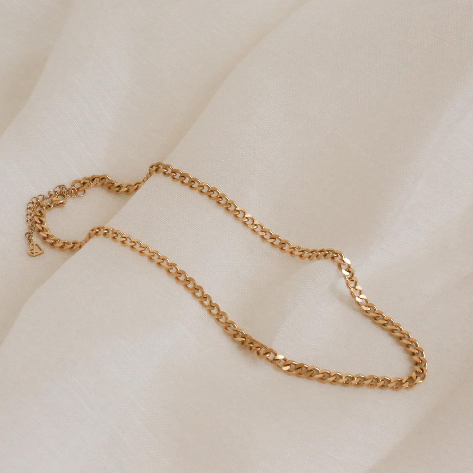 A close-up shot of a chunky gold curb chain necklace with flat, interlocking links, draped gently across a soft ivory-colored fabric