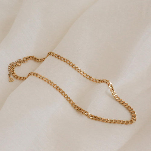 A close-up shot of a chunky gold curb chain necklace with flat, interlocking links, draped gently across a soft ivory-colored fabric