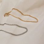 A flat lay showing both the gold and silver versions of the curb chain necklace on a cream fabric.