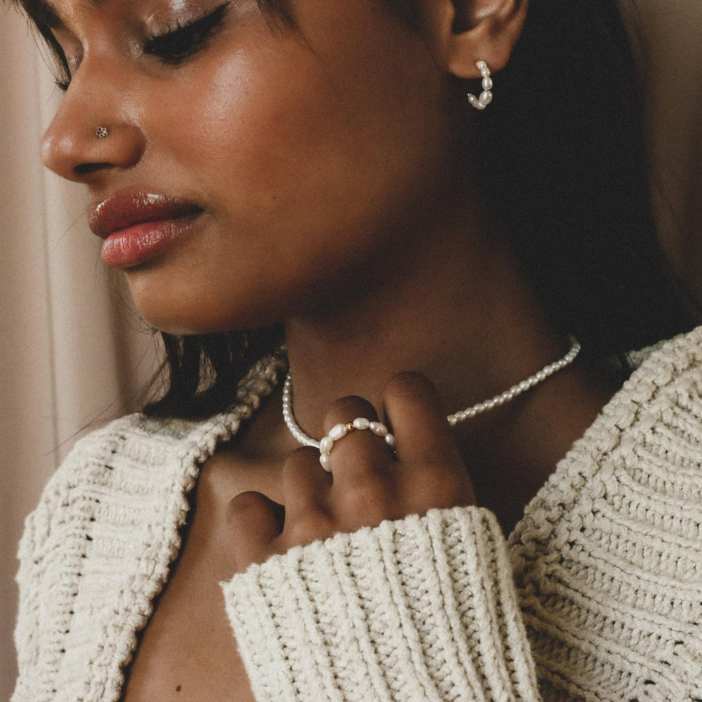A woman in a cream knit sweater, wearing freshwater pearl earrings, necklace, and the Pearl Beaded Ring, creates a classy accessory ensemble as she gazes downward.