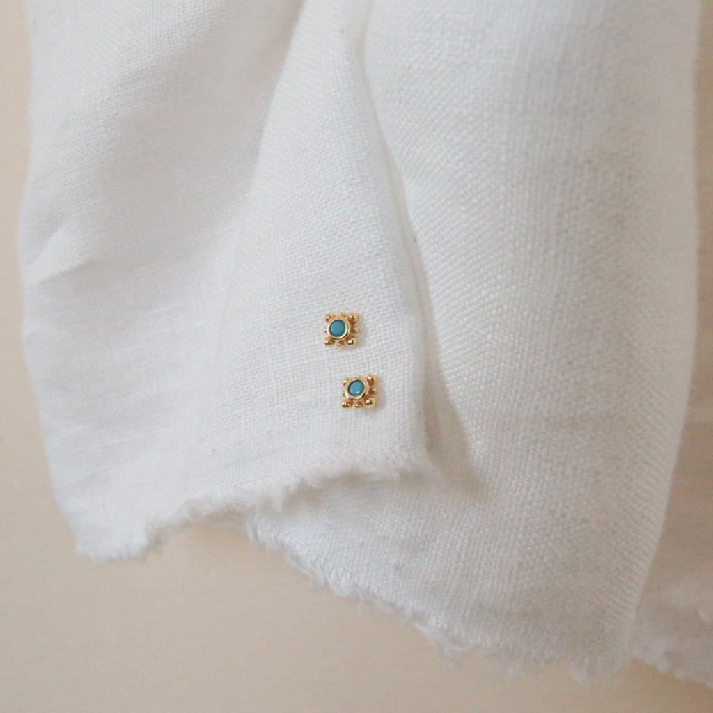 Two small square gold Mara Studs with turquoise stone centers rest on a textured white fabric.