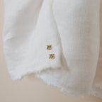 Two small square gold Mara Studs with turquoise stone centers rest on a textured white fabric.