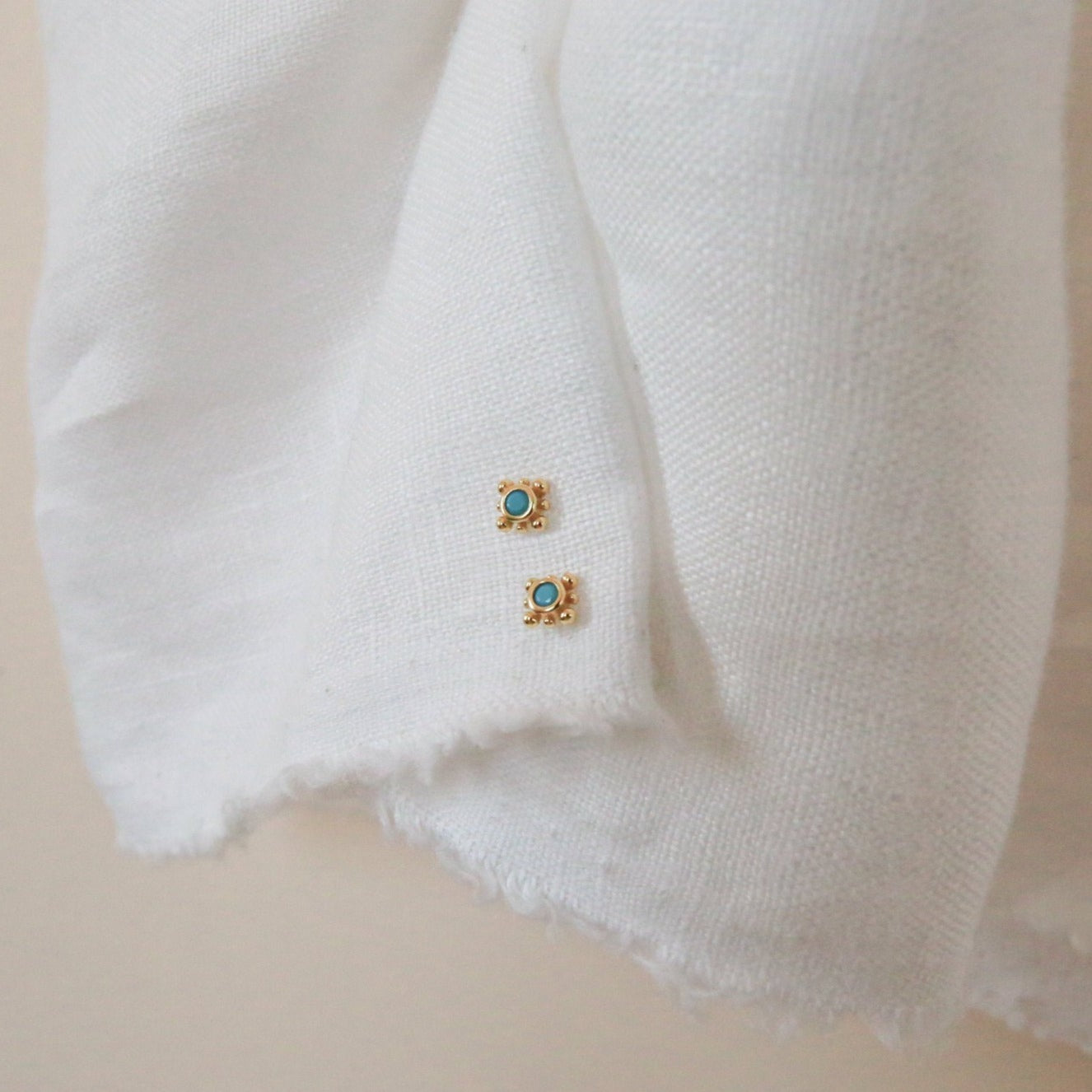 Two small square gold Mara Studs with turquoise stone centers rest on a textured white fabric.