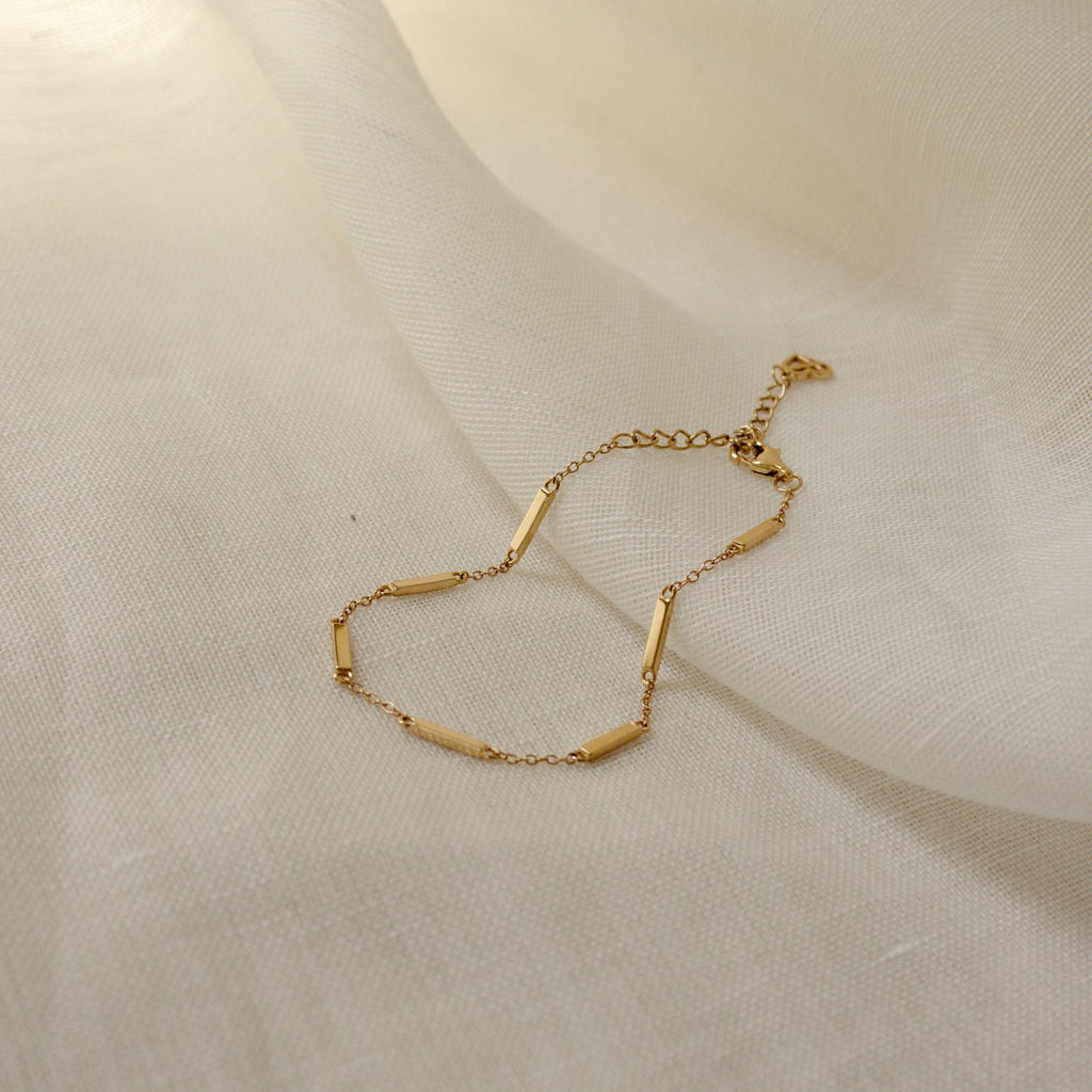 The Abby Bar Link Bracelet is a delicate gold satellite bracelet with small rectangular bar accents, shown on a lightly textured fabric background.