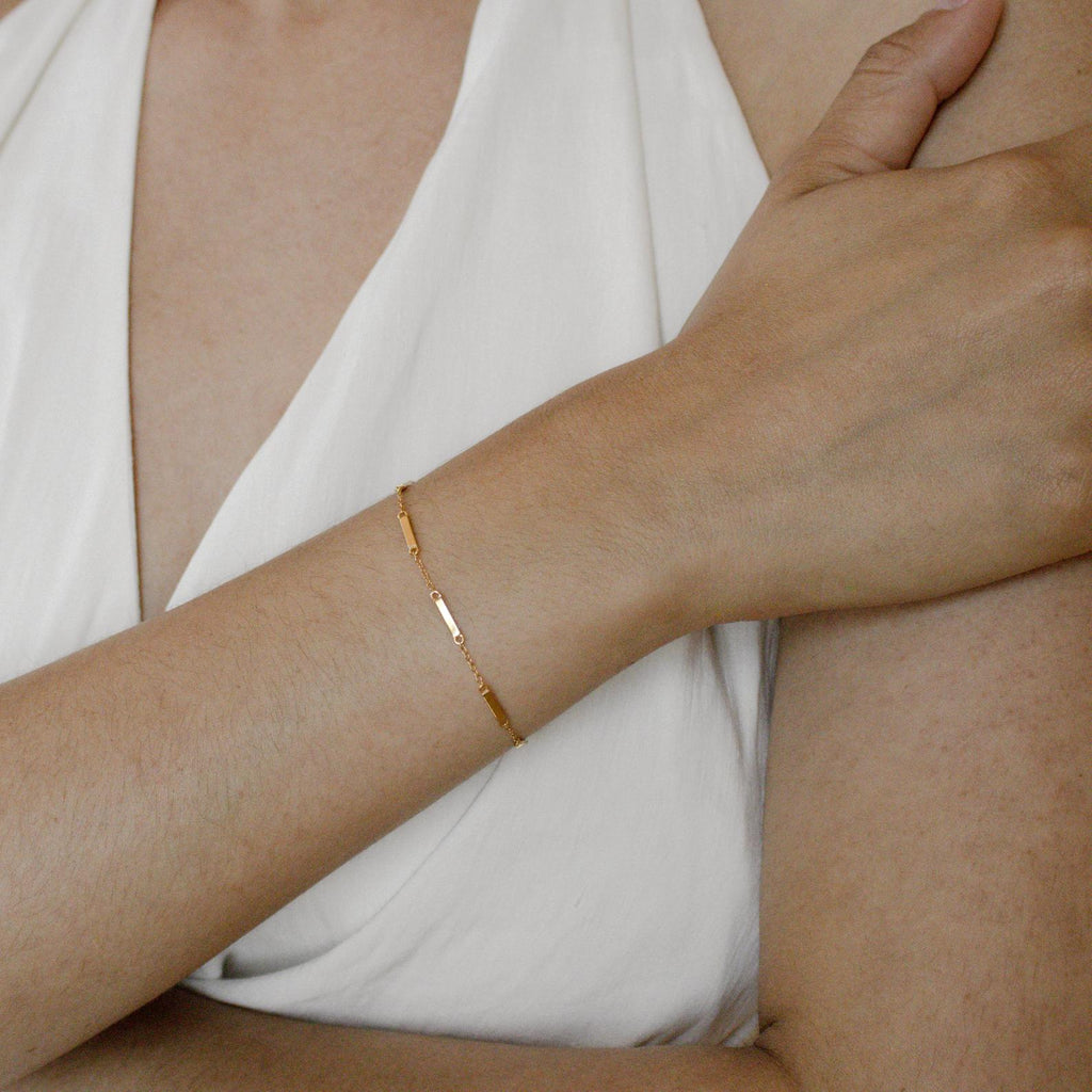 A person wearing a sleeveless white top showcases the Abby Bar Link Bracelet, a dainty adjustable accessory adorning their wrist as they rest their arm across their body.