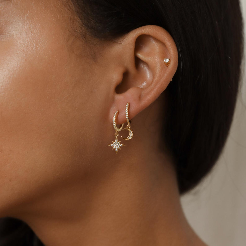 Close-up of a woman's ear adorned with Moon and Star Huggies in 18K Gold, featuring three constellation-inspired gold earrings—a small stud, a hoop, and a star-shaped dangle.