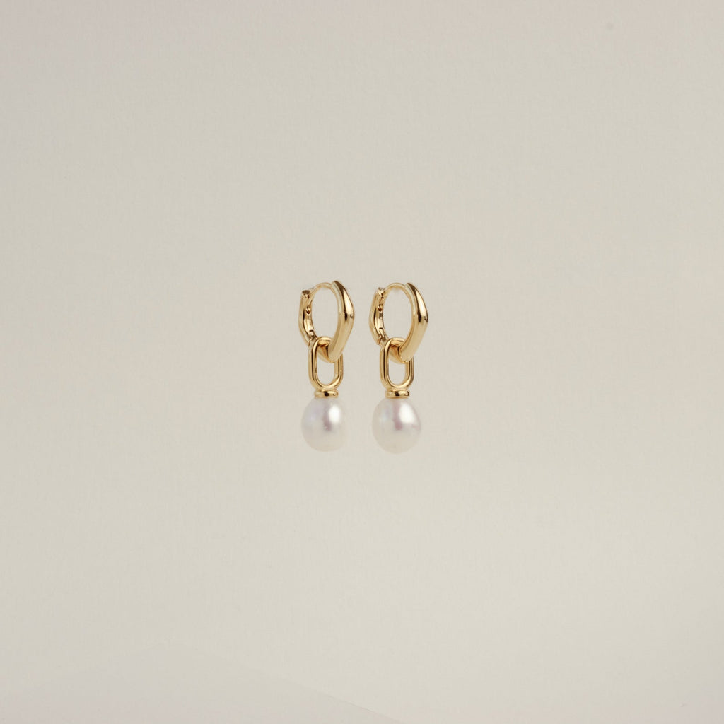 The Hazel Pearl Hoops are gold hoop earrings with dangling white pearls, a modern jewelry standout, elegantly showcased against a plain light background.