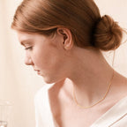 A woman with light brown hair in a low bun wears gold Dainty Bar Studs and a white top, looking down at a glass.